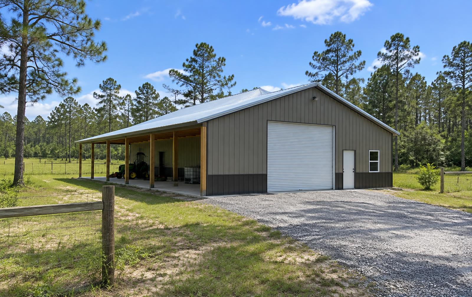 Metal-sided post-frame pole barn with roll-up door and open lean-to on pasture land with pines